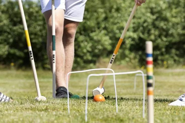 Untere Beine einer Person in Shorts und Turnschuhen auf Rasen, mehrere Schl&auml;ger mit langen Stielen, ein orangefarbener Ball, metallische Torb&ouml;gen und ein Holzpfahl mit farbigen Ringen