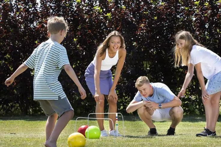 Vier Personen auf Rasen um mehrere bunte B&auml;lle (gelb, rot, gr&uuml;n) und ein niedriges Drahtgestell; im Hintergrund eine dichte Hecke