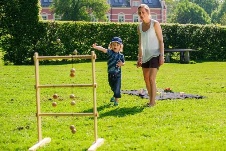 Erwachsene Person und Kind auf Wiese; Kind wirft Ball zu Holzrahmen mit aufgeh&auml;ngten Kugeln; Decke auf Gras