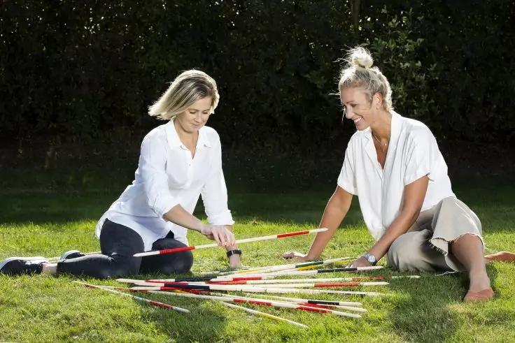 Zwei Personen sitzen auf dem Rasen und legen lange St&auml;be mit roten, gelben und schwarzen Markierungen