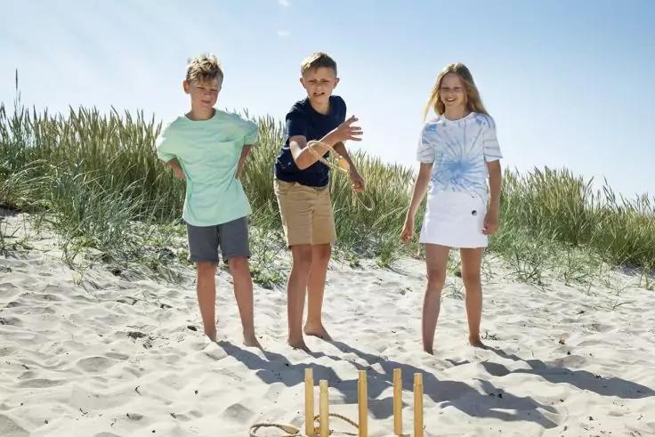 Drei Kinder stehen auf einem Sandstrand vor D&uuml;nen mit Gras; im Vordergrund liegt ein h&ouml;lzernes Wurfspiel im Sand