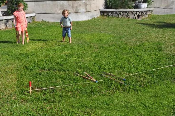 Zwei Kinder stehen auf einem Rasen; im Vordergrund liegen mehrere Holzst&ouml;cke und ein gespanntes Seil auf dem Gras; im Hintergrund eine niedrige Steinmauer mit Pflanzen