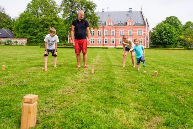 Vier Personen auf einer Rasenfl&auml;che mit h&ouml;lzernen Kl&ouml;tzen und St&auml;ben; im Hintergrund rotes Backsteingeb&auml;ude und B&auml;ume
