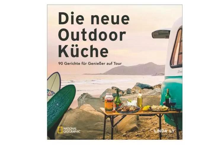 Buchcover mit Titel 'Die neue Outdoor K&uuml;che &mdash; 90 Gerichte f&uuml;r Genie&szlig;er auf Tour'. Strandszene mit zwei Surfboards, einem Campingtisch mit Gl&auml;sern und Tellern, Felsstrand, Meer, Bergen im Hintergrund und einem t&uuml;rkisen Wohnmobil am rechten Bildrand