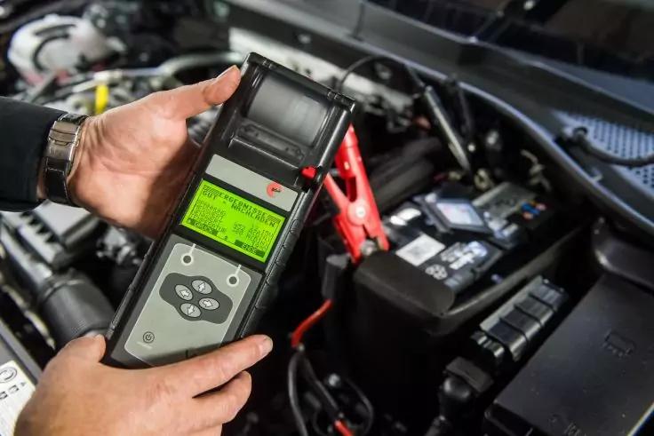 H&auml;nde halten ein Handger&auml;t mit gr&uuml;n beleuchtetem Display und Tasten; rote Krokodilklemme an einem Batteriepol im offenen Motorraum befestigt