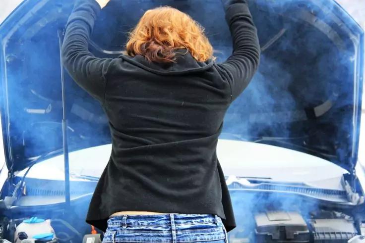 Person von hinten mit roten Haaren, schwarzer Jacke und blauer Jeans, h&auml;lt die ge&ouml;ffnete Motorhaube eines Autos hoch, Rauch steigt aus dem Motorraum