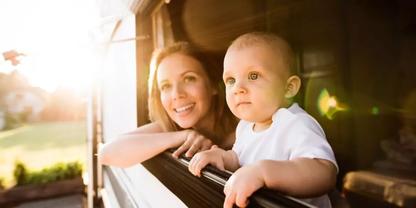 Frau und Baby lehnen an offenem Fensterrahmen aus Holz, Sonnenlicht f&auml;llt von links