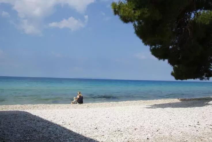 Person sitzt am kieseligen Strand am Ufer, t&uuml;rkisblaues Meer bis zum Horizont, Baum rechts, Schatten im Vordergrund, teilweise bew&ouml;lkter Himmel