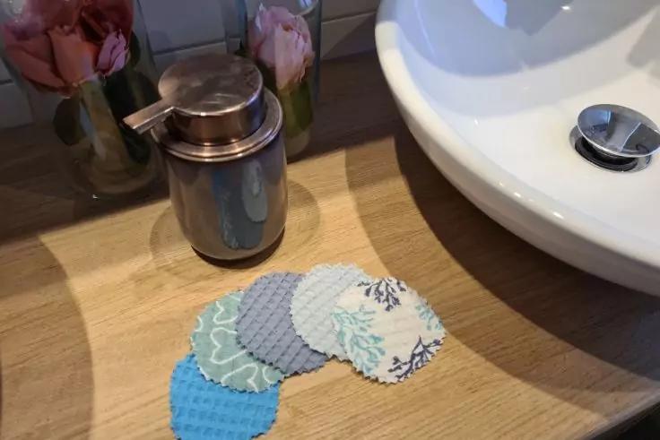 Waschbecken auf Holzablage, mehrere runde gemusterte Stoffpads, kleiner Metallbehälter, rosa Rosen im Glas