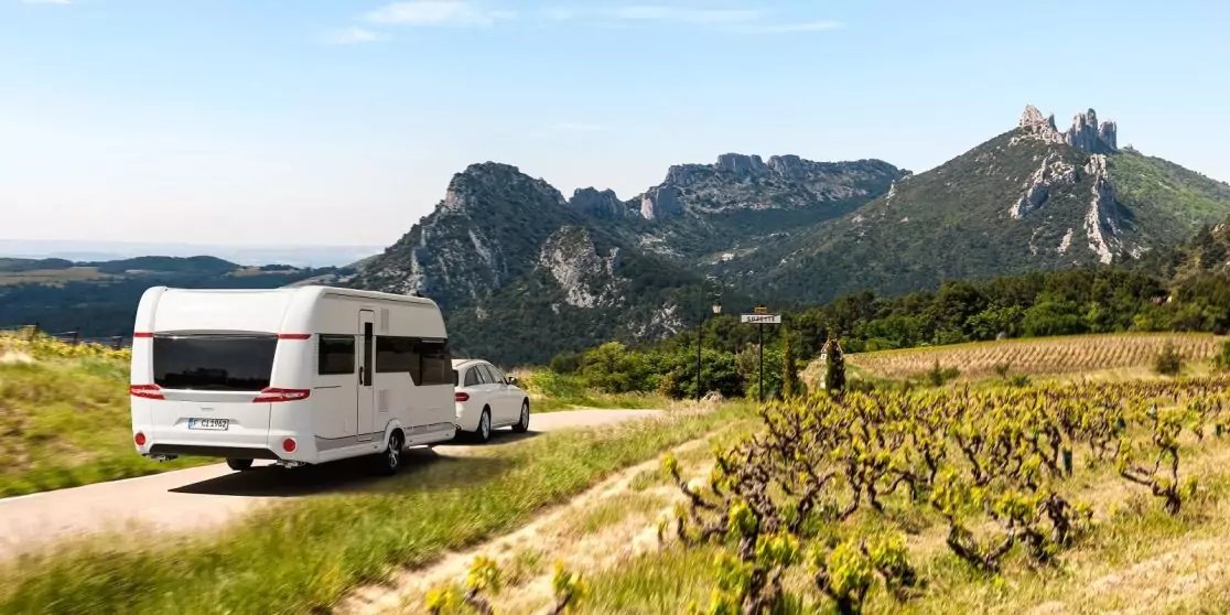Wei&szlig;es Wohnmobil und wei&szlig;es Auto auf Landstra&szlig;e neben Rebreihen; Felsformationen und ein steinernes Geb&auml;ude auf einem H&uuml;gel; blauer Himmel