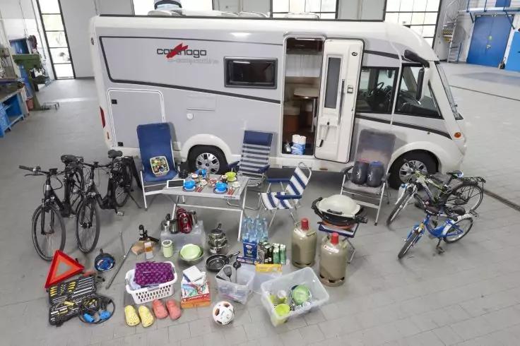 Wei&szlig;es Wohnmobil in einer Halle, davor mehrere Fahrr&auml;der und auf dem Boden angeordnete Gegenst&auml;nde: Klappst&uuml;hle, kleiner Tisch, Gaskocher und Gasflaschen, K&uuml;hlboxen, Kisten und Taschen, Warn-Dreieck, Schuhe und ein Ball