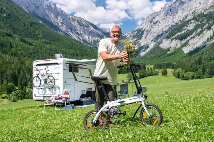 Person steht auf gr&uuml;ner Wiese neben einem Klapprad und h&auml;lt einen Blumenstrau&szlig;; im Hintergrund ein Wohnmobil mit zwei Fahrr&auml;dern an der Heckklappe, ein Campingtisch und ein Stuhl, Berge und bew&ouml;lkter Himmel