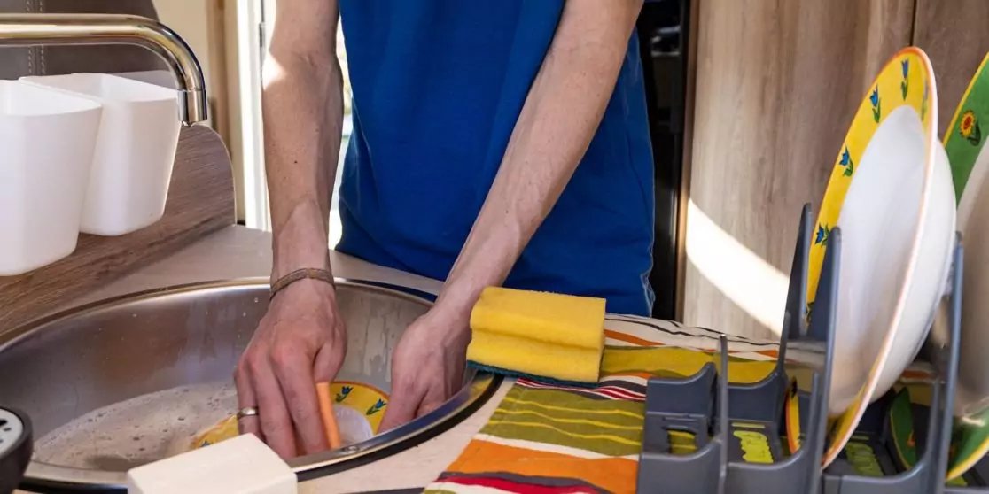 H&auml;nde und Unterarme einer Person in blauem T-Shirt an einem Sp&uuml;lbecken, gelber Schwamm, Geschirrkorb mit Tellern und Besteck