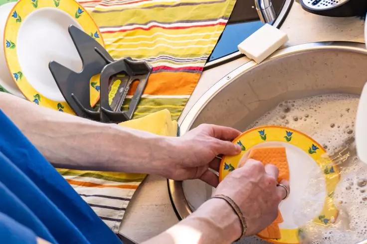 H&auml;nde sp&uuml;len einen gelben Teller mit gr&uuml;nen Mustern in einem Sp&uuml;lbecken, schaumiges Wasser, Schwamm und gestreiftes Geschirrtuch sichtbar