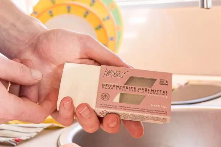 Zwei H&auml;nde halten eine kleine rechteckige Kartonbox mit aufgedrucktem Text und einem transparenten Sichtfenster &uuml;ber einem Sp&uuml;lbecken