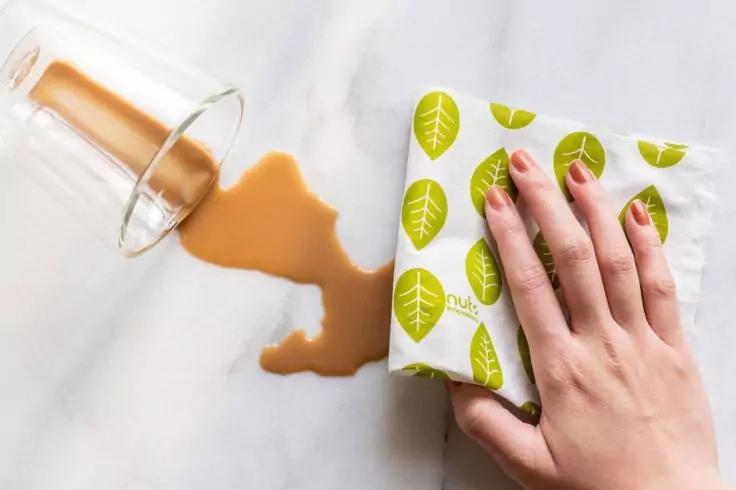 Hand mit lackierten Fingern&auml;geln h&auml;lt ein wei&szlig;es Tuch mit gr&uuml;nem Blattmuster auf einer wei&szlig;en Oberfl&auml;che; daneben ein umgekipptes Glas und eine versch&uuml;ttete braune Fl&uuml;ssigkeit