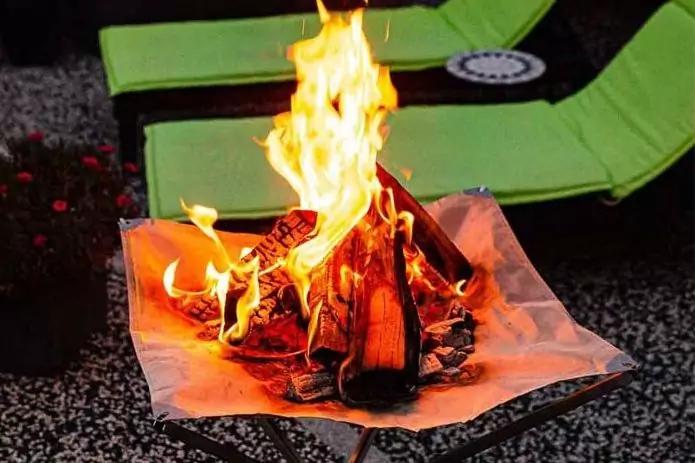 Brennender Holzstapel in einer flachen Metallschale auf einem Gestell, mit heller Unterlage darunter, im Hintergrund zwei gr&uuml;ne Liegest&uuml;hle auf Kies, links rote Blumen