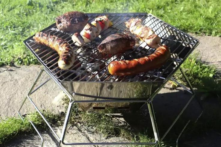 Tragbarer Metallgrill mit mehreren W&uuml;rstchen und Gem&uuml;sespie&szlig;en auf dem Rost, Gras und Gehweg im Hintergrund