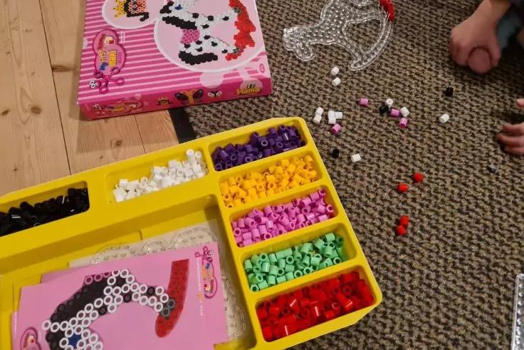 Gelber Sortierkasten mit farbigen B&uuml;gelperlen in mehreren F&auml;chern; rosa Steckplatte mit Perlen darauf; verstreute Perlen und eine Perlenkette auf grauem Teppich; rosa Karton mit Hundemotiv auf Holzboden; H&auml;nde am Bildrand