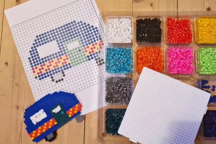 Blatt mit Punktvorlage einer bunten pixelartigen Form, mehrere Kunststofff&auml;cher mit verschiedenfarbigen B&uuml;gelperlen, fertiges blau-orangetes Perlenobjekt und wei&szlig;e Steckplatte auf Holzuntergrund