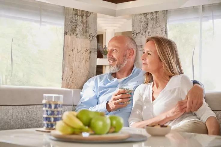 Ein Mann mit grauem Bart und hellblauem Hemd und eine Frau mit langen blonden Haaren und wei&szlig;er Bluse sitzen nebeneinander auf einem Sofa; der Mann hat den Arm um die Frau gelegt. Auf dem Tisch davor eine Schale mit gr&uuml;nen &Auml;pfeln und Bananen sowie ei