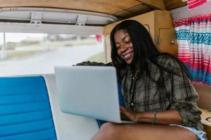 L&auml;chelnde Person sitzt in einem Van mit offenem Laptop auf dem Scho&szlig;, tr&auml;gt kariertes Hemd und kurze Hose, bunte Decke an der Seitenwand