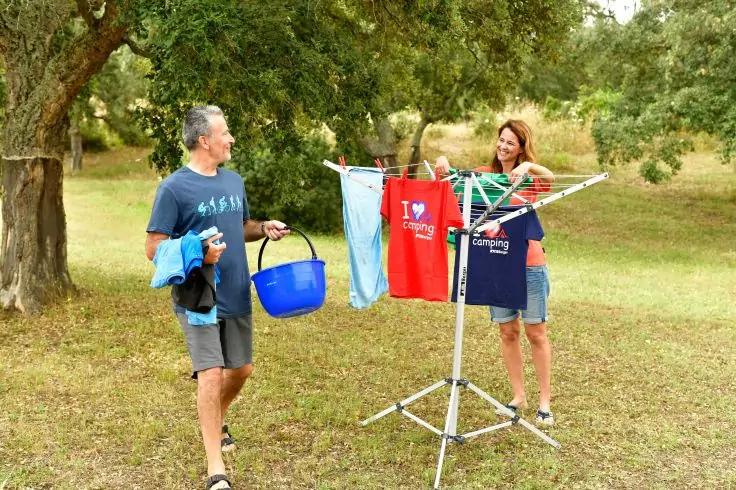 Mann und Frau h&auml;ngen W&auml;sche an klappbarem W&auml;schest&auml;nder auf einer Wiese mit B&auml;umen. Mann h&auml;lt blauen Eimer; sichtbare Kleidungsst&uuml;cke: rotes T-Shirt mit Aufschrift 'I &hearts; camping' und blaues T-Shirt