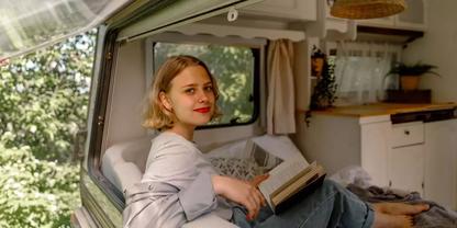 Person sitzt in der T&uuml;r&ouml;ffnung eines Wohnmobils, h&auml;lt ein offenes Buch und blickt zur Kamera; kurzes blondes Haar, rote Lippen; Vorhang, Regal mit Pflanze und B&auml;ume drau&szlig;en sichtbar