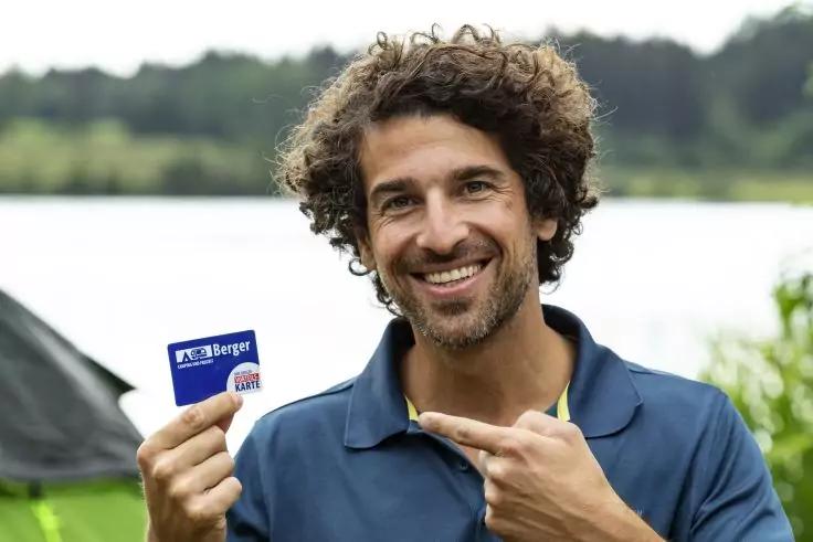 Person mit lockigen dunklen Haaren, l&auml;chelnd, h&auml;lt eine blaue Karte und zeigt mit einer Hand darauf, tr&auml;gt dunkelblaues Poloshirt, im Hintergrund See und Zelt