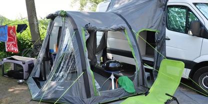 Graues Vorzelt mit transparenten Fenstern, an einem wei&szlig;en Wohnmobil befestigt; gr&uuml;ner Klappstuhl im Vordergrund, runder Grill auf Stativ im Vorzelt, lila Transportbox und aufgeh&auml;ngte Handt&uuml;cher links; Grasboden und B&auml;ume im Hintergrund