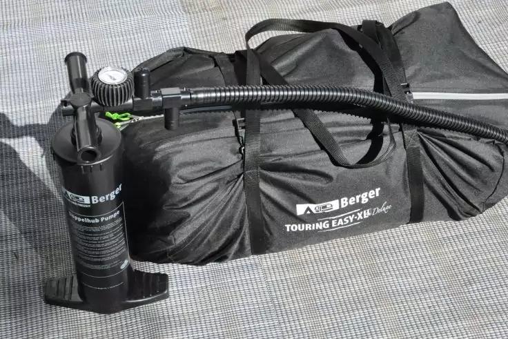 Schwarze Handpumpe mit Manometer und Schlauch, verbunden mit einer zusammengelegten schwarzen Tasche mit Tragegriffen und weißem Aufdruck auf einer grauen Matte