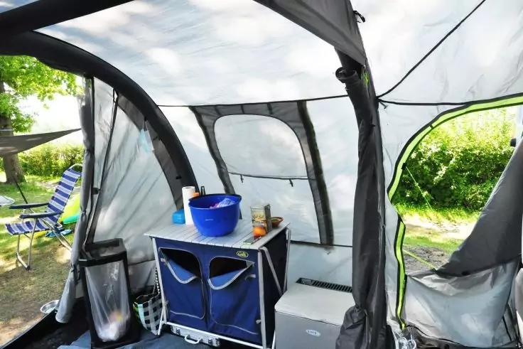 Innenraum eines grauen Stoffzelts mit gebogenem Gestänge und Netzfenstern, blaues Aufbewahrungsmöbel mit zwei Stoffschubladen und blauer Wanne darauf, kleine weiße Kühlbox, Mülleimer und gestreifter Klappstuhl außen auf Gras