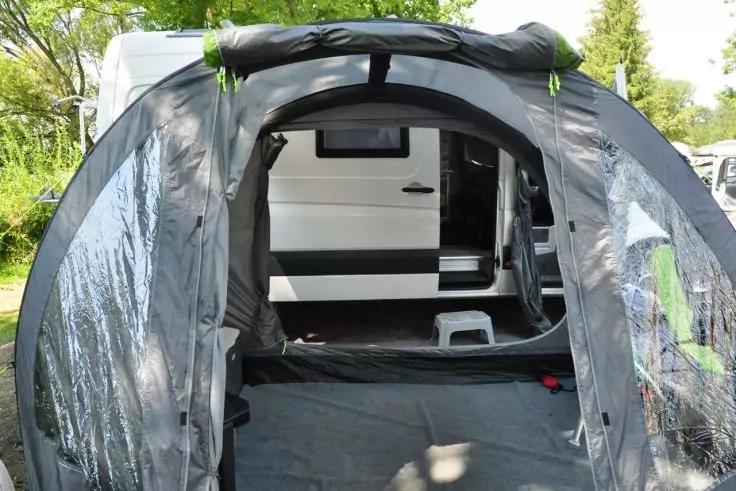 Blick aus dem Inneren eines grauen Zeltvorbaus mit transparenten Fenstern auf die Seite eines weißen Wohnmobils mit geöffneter Tür und einem weißen Tritthocker, Schatten von Bäumen auf dem Boden