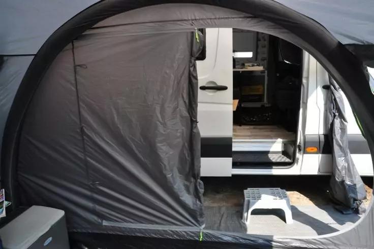 Blick durch den Eingang eines Zelts auf die offene Seitentür eines weißen Wohnmobils und eine weiße Trittstufe