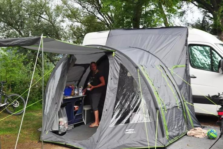 Graues Vorzelt an einem weißen Kastenwagen auf einer Wiese; eine Person steht im Vorzelt neben einem Tisch mit Flaschen und Utensilien; Fahrrad lehnt an einem Baum, Bäume im Hintergrund