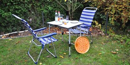 Zwei blau-wei&szlig; gestreifte Klappst&uuml;hle und ein kleiner wei&szlig;er Klapptisch auf Rasen; auf dem Tisch zwei Flaschen, zwei Gl&auml;ser und ein Teller; runder orangefarbener, gelochter Gegenstand lehnt am Tisch; Hecke im Hintergrund, verstreute Bl&auml;tter