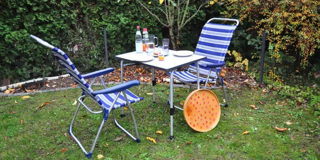 Zwei blau-wei&szlig; gestreifte Klappst&uuml;hle und ein kleiner wei&szlig;er Klapptisch auf Rasen; auf dem Tisch zwei Flaschen, zwei Gl&auml;ser und ein Teller; runder orangefarbener, gelochter Gegenstand lehnt am Tisch; Hecke im Hintergrund, verstreute Bl&auml;tter