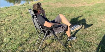 Person in Klappstuhl auf Gras, h&auml;lt Telefon ans Ohr, Wasser im Hintergrund