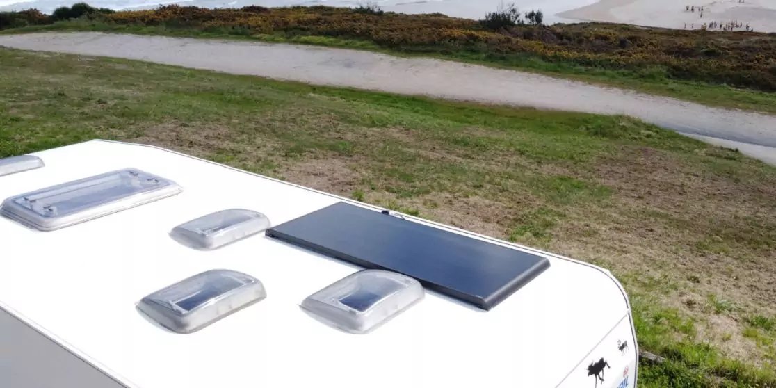 Dach eines wei&szlig;en Wohnwagens mit vier wei&szlig;en Dachluken und einem schwarzen rechteckigen Panel, im Hintergrund Gras, ein schmaler Wassergraben und sandige D&uuml;nen