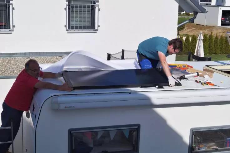 Zwei Personen auf dem Dach eines wei&szlig;en Wohnmobils, mit Werkzeugen daneben; im Hintergrund eine Hausfassade mit zwei Fenstern