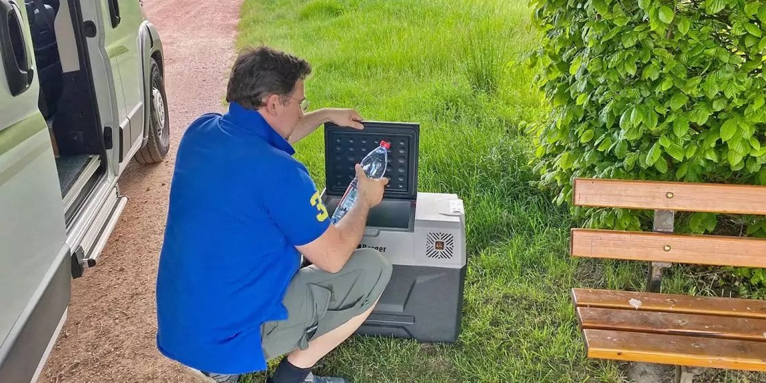 Person kniet neben einem offenen wei&szlig;en Transporter und ber&uuml;hrt ein tragbares Ger&auml;t auf dem Boden neben einer Holzbank und einem gr&uuml;nen Busch