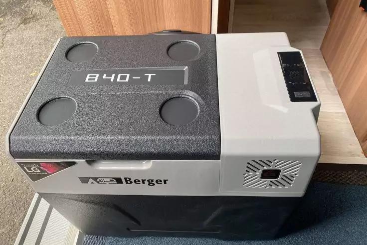 Graue Box mit Aufschriften "Berger" und "840-T", schwarzer Deckel mit vier runden Vertiefungen, Bedienfeld und L&uuml;ftungs&ouml;ffnung rechts, steht auf blauem Boden vor einem Holzschrank