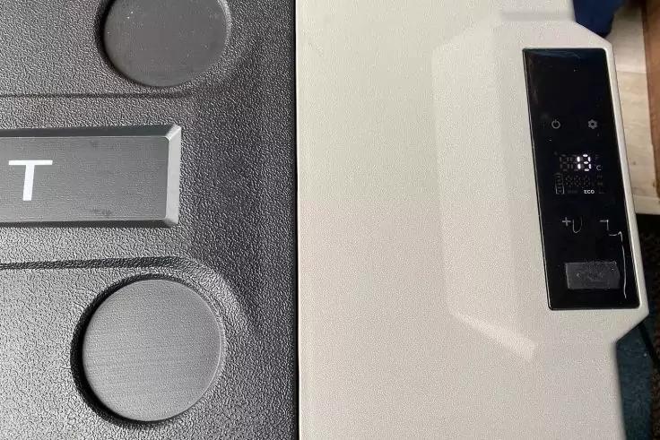 Nahaufnahme eines Ger&auml;ts: links schwarzer, strukturierter Kunststoff mit zwei runden Vertiefungen und grauem Schild mit dem Buchstaben 'T'; rechts hellgraue Fl&auml;che mit schwarzem Bedienfeld mit Symbolen und Tasten; blauer Stoff am rechten unteren Rand