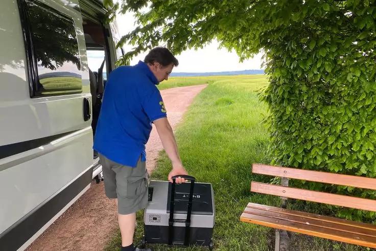 Person in blauem T-Shirt und gr&uuml;nen Shorts schiebt eine rollbare schwarz-graue Box mit Teleskopgriff neben einem wei&szlig;en Wohnmobil; rechts eine Holzbank, im Hintergrund Grasweg und Felder unter B&auml;umen
