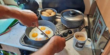 Zwei Eier in einer Pfanne auf dem Herd; H&auml;nde halten Pfanne und Pfannenwender; Schale mit Eiern, zwei T&ouml;pfe mit Deckel, Kaffeetasse und ein aufgestelltes Tablet auf der Arbeitsfl&auml;che