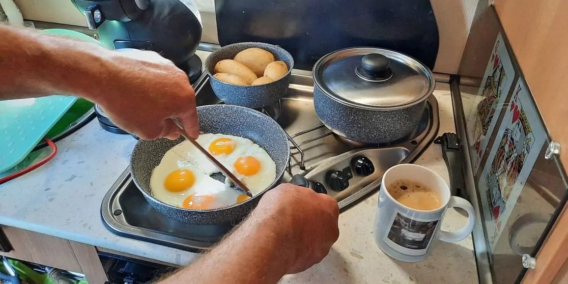 Zwei Eier in einer Pfanne auf dem Herd; H&auml;nde halten Pfanne und Pfannenwender; Schale mit Eiern, zwei T&ouml;pfe mit Deckel, Kaffeetasse und ein aufgestelltes Tablet auf der Arbeitsfl&auml;che