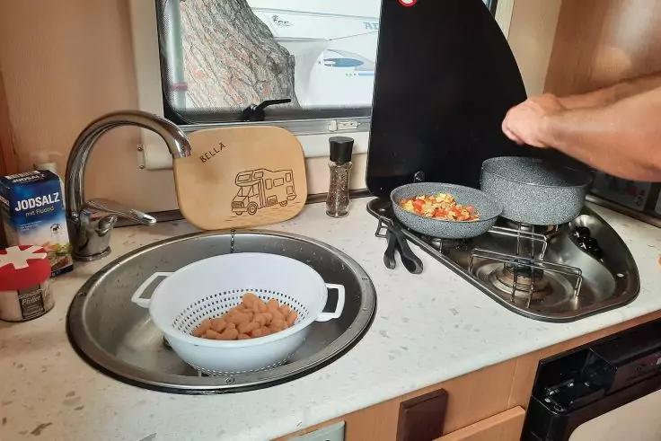 Kleine K&uuml;chenzeile: Sp&uuml;le mit Abtropfsieb mit braunem Inhalt, Wasserhahn, Schneidebrett mit Motiv, Salzpackung, Gew&uuml;rzm&uuml;hle, Gasherd mit Pfanne und Topf; Hand h&auml;lt einen Deckel; Fenster mit Baum im Hintergrund
