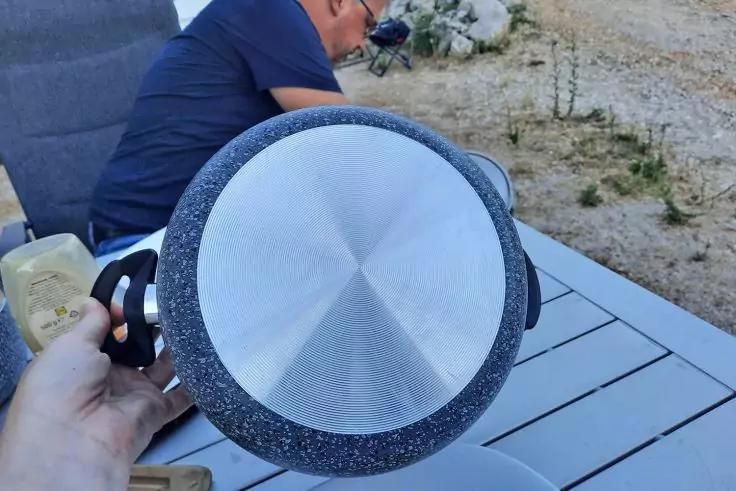 Hand h&auml;lt Bratpfanne mit sichtbarem Metallboden und schwarzem Rand; im Hintergrund Tisch, Teller und sitzende Person im Freien