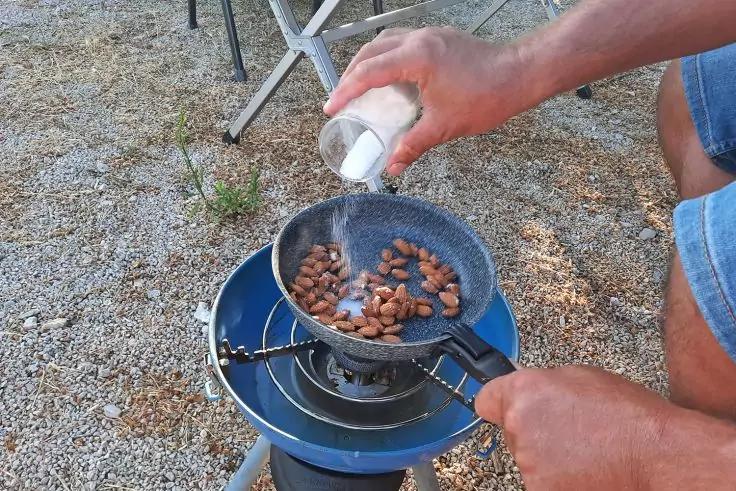 H&auml;nde gie&szlig;en wei&szlig;e K&ouml;rner aus einem kleinen Beh&auml;lter in eine Pfanne mit braunen Mandeln auf einem blauen Campingkocher; Person tr&auml;gt blaue Shorts; Kiesboden und Klappstuhl im Hintergrund