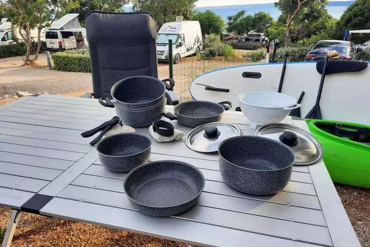 Mehrere T&ouml;pfe, Pfannen, Sch&uuml;sseln und Deckel auf einem Au&szlig;entisch; im Hintergrund Campingstuhl, Transporter, wei&szlig;es Paddleboard, gr&uuml;nes Kajak, B&auml;ume und Wasser